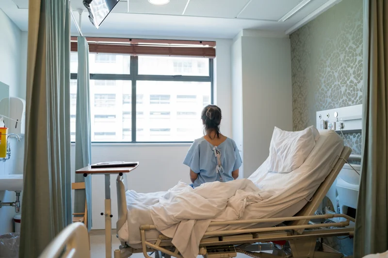A woman sits on a hospital bed and is looking out a window.