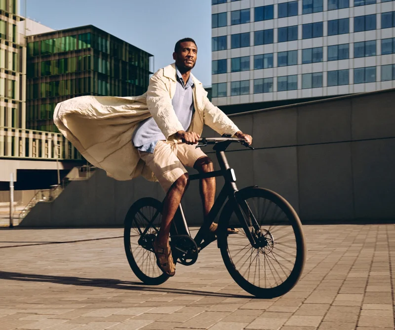 A photograph. A man riding a Cowboy e-bike through an urban area, with his coat flowing dramatically behind him.