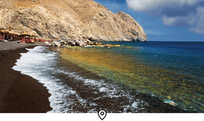 Image Description: Gentle waves crashing onto Perissa Beach, a famous black sand beach in Santorini, with travelers relaxing on sun loungers in the background