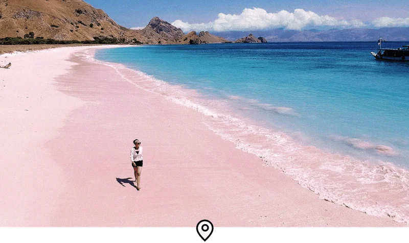 Image Description: A female traveler strolling barefoot along Indonesia’s iconic Pink Beach, flanked by turquoise waters