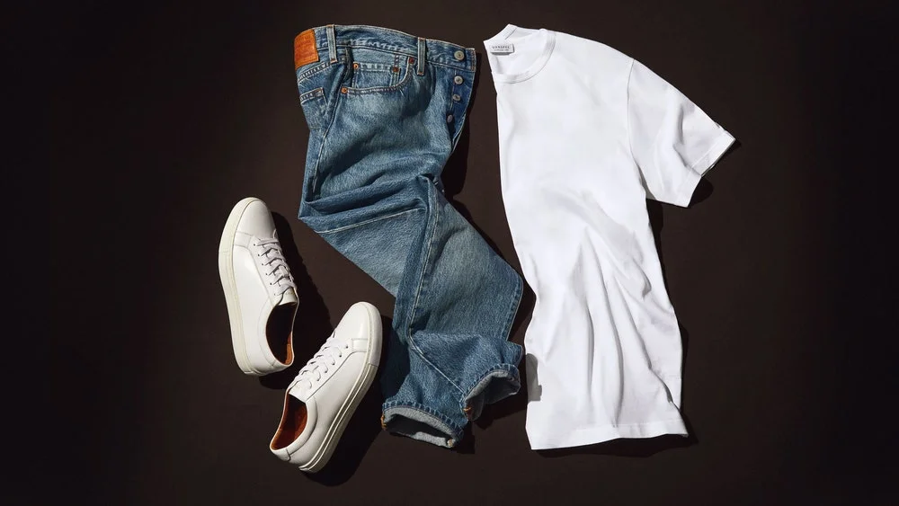 Image of a white t shirt, light jeans, and white leather sneakers laid out on a dark grey background