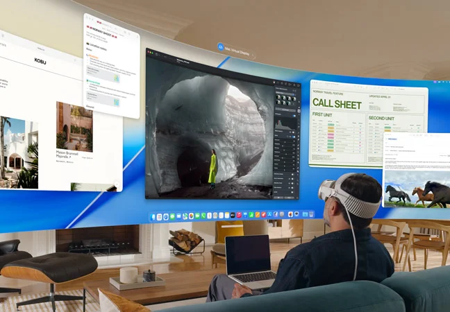 Person wearing Apple Vision Pro sits on a couch in a living room with an open MacBook on lap, ultrawide screen Mac Virtual Display floats in front of the person, filling the room