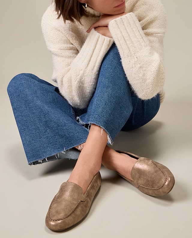 A woman sitting cross-legged in a cozy off-white turtleneck sweater, raw-edge jeans, and the Ozzie Loafer in Bronze. Shop Now