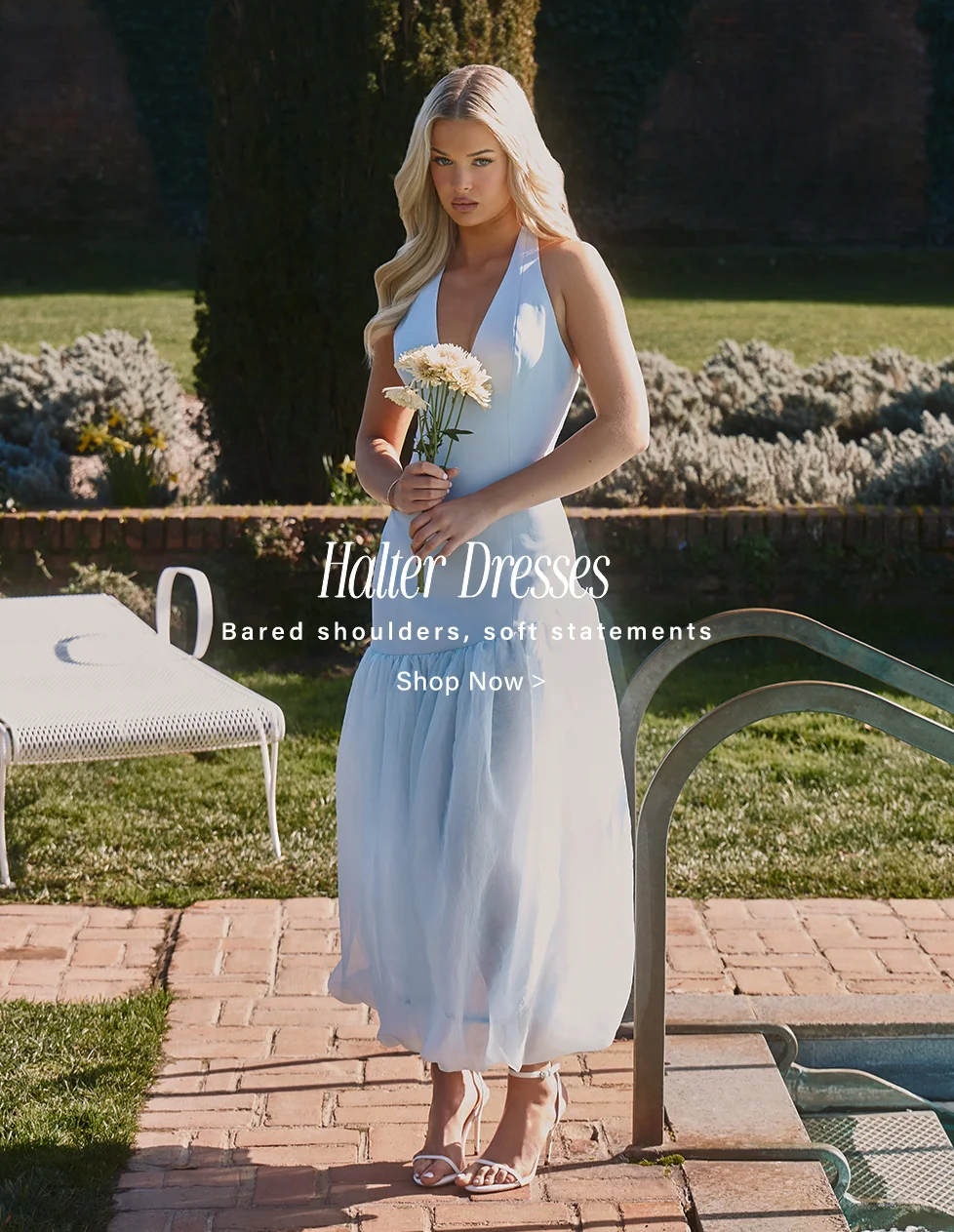 Blonde woman in a flowing light blue halter dress and white heels, holding white flowers, stands outdoors by a pool. Shop our chic halter dresses.