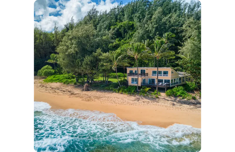 An expansive yellow home is situated on a pristine beach in Hawaii. It is set back among the palm trees and lush greenery with nothing else in sight except for the blue sea lapping the shore.