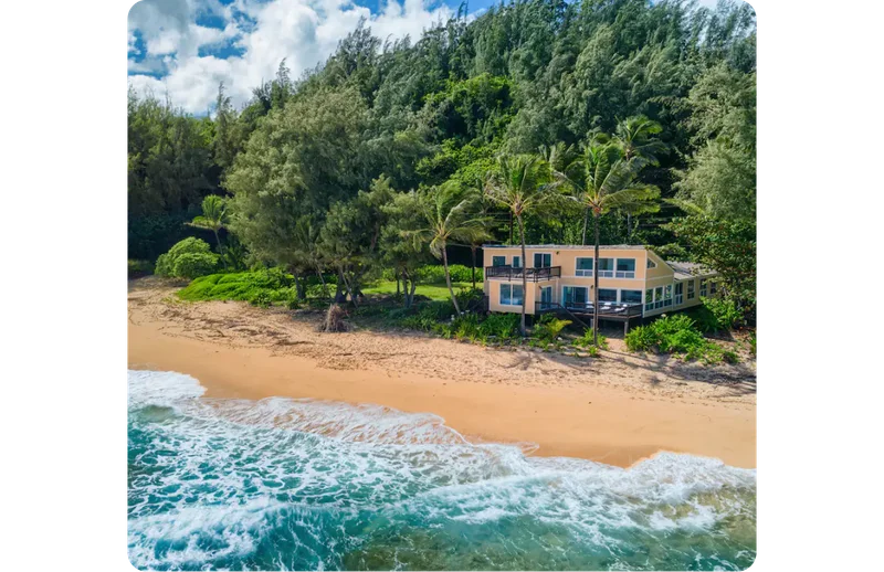 An expansive yellow home is situated on a pristine beach in Hawaii. It is set back among the palm trees and lush greenery with nothing else in sight except for the blue sea lapping the shore.