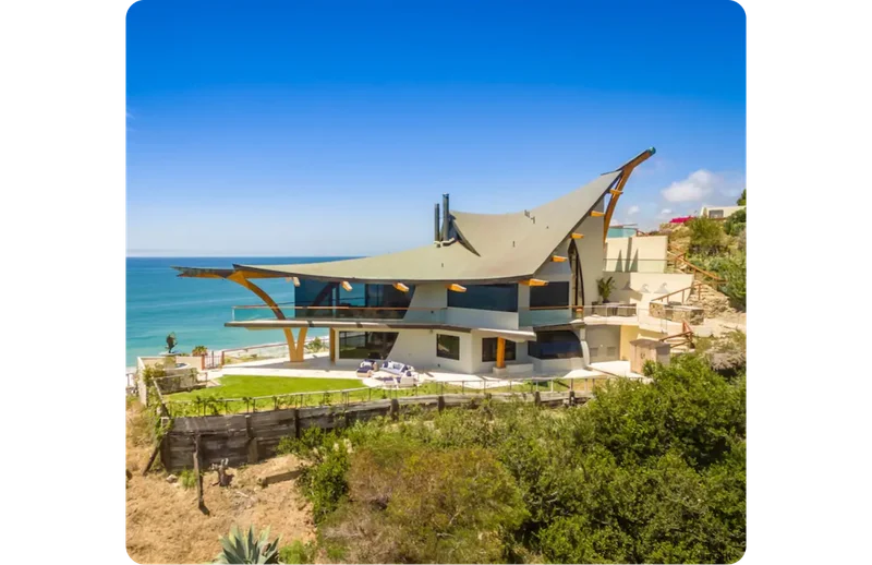 An image of Eagle’s Watch, an expansive home overlooking the Pacific Ocean designed by Harry Gesner. The triangular roofline curves upward with a pointed beam, and big glass windows offer panoramic views of the beach. An open-air porch wraps around the house set above a green lawn.