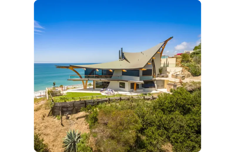 An image of Eagle’s Watch, an expansive home overlooking the Pacific Ocean designed by Harry Gesner. The triangular roofline curves upward with a pointed beam, and big glass windows offer panoramic views of the beach. An open-air porch wraps around the house set above a green lawn.
