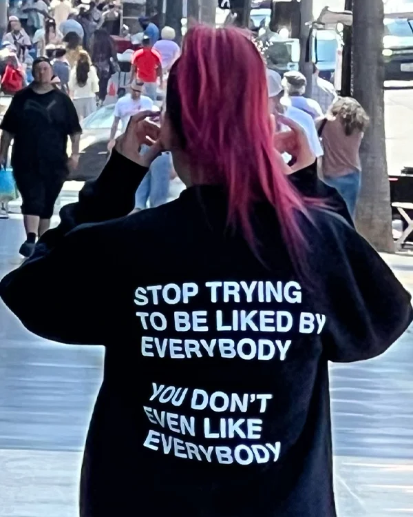 image of a woman walking down the city streets with link hair in a ponytail she is wearing the We're Not Really Strangers Stop Trying Crewneck. It is black and in white writing it says "STOP TRYING TO BE LIKED BY EVERYBODY YOU DON'T EVEN LIKE EVERYBODY"