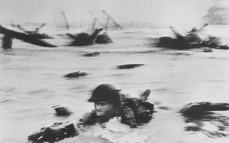 A black and white photo of an American soldier crawling in the surf as the Allied forces storm the beaches of Normandy on D-Day.
