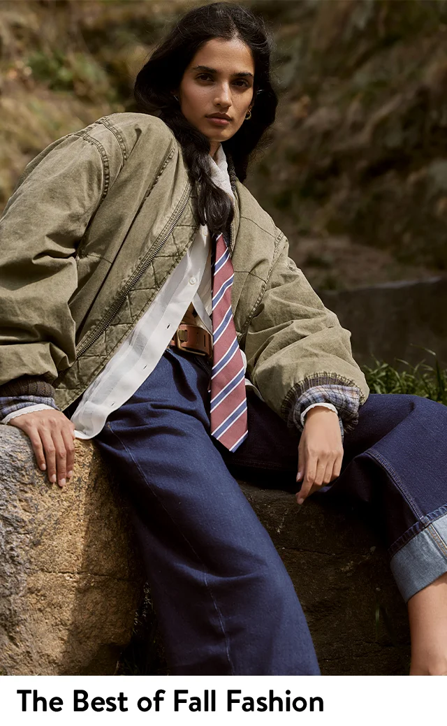 A woman wearing a bomber jacket, button-up shirt, tie and jeans.