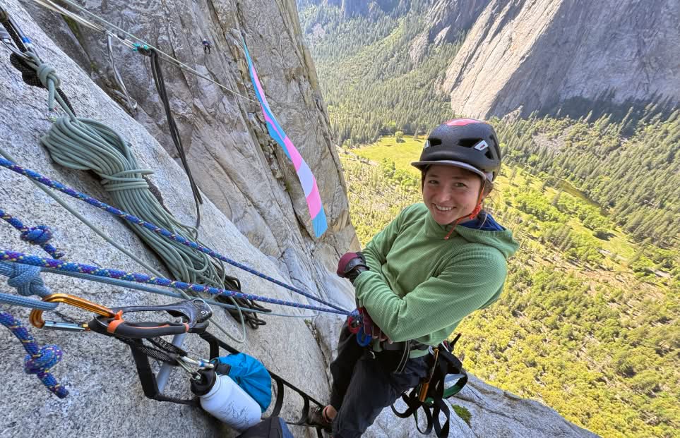 A climber anchored high on a rock wall with a colorful flag flying in the background.