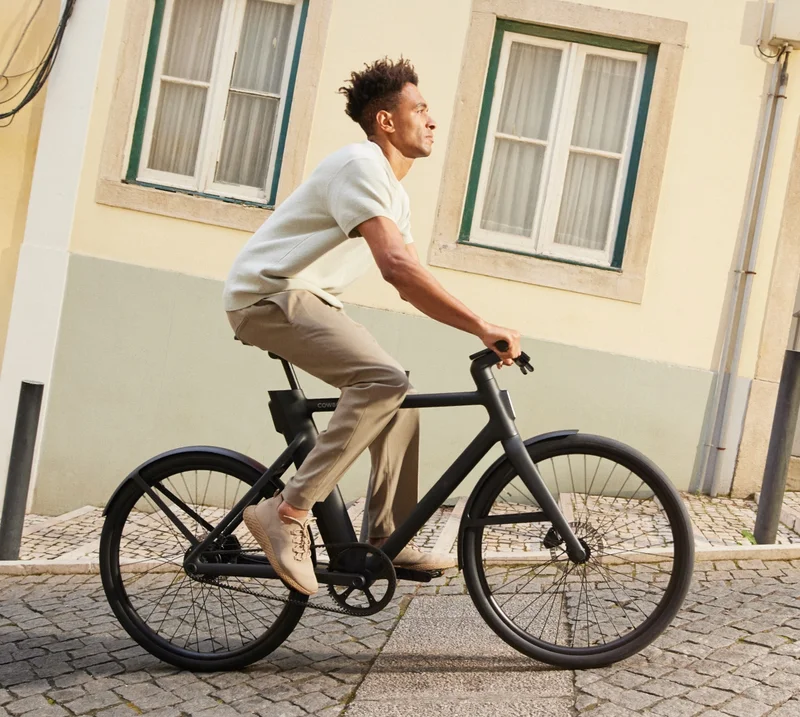 A man riding a sleek black Cowboy bike on what appears to be a flat cobblestone street in a sunny urban neighbourhood. The camera angle cleverly conceals the steep incline of the hill, creating the illusion of effortless riding in a picturesque setting framed by pastel-coloured buildings.