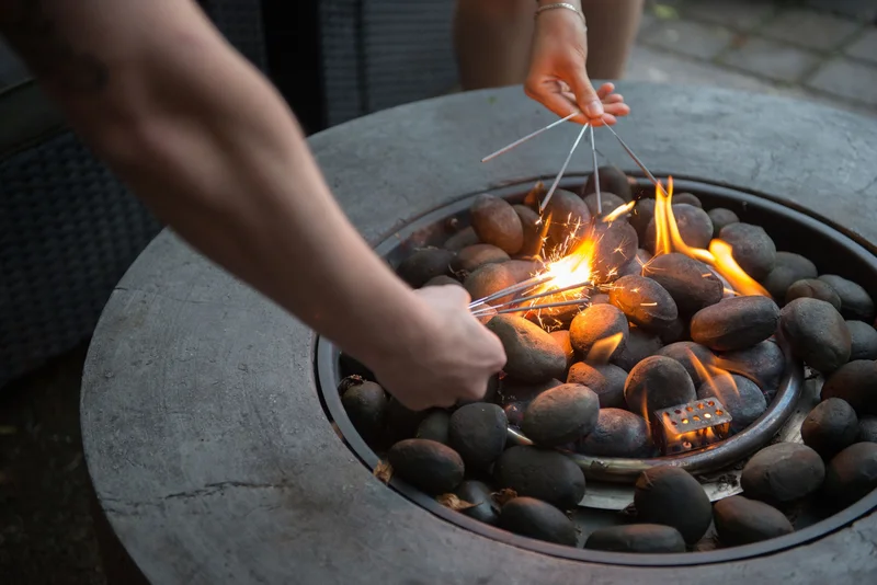Two people reach arms over top a round grey fire pit to light sparklers.