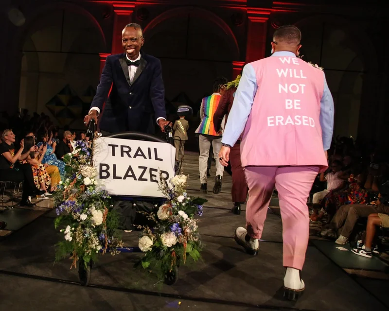 Models walk on a runway; one has a wheelchair adorned in flowers that says “TRAIL BLAZER” and another has a pink vest that says “WE WILL NOT BE ERASED”