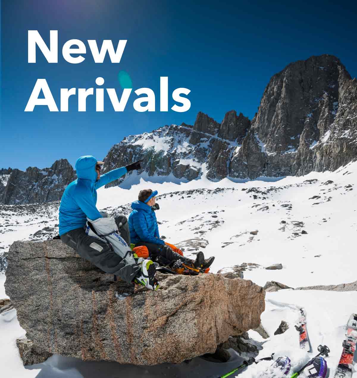 New Arrivals. Two backcountry skiers sit on a rock and point up at the mountain high above them.