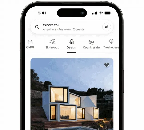 An animated gif of a phone opens to the categories section of the Airbnb app. It cycles between categories, showing different homes. We start on the Design Category with a home that looks as if it has been built with white blocks. Large open windows show light coming from inside. It slides to the left and we’re on the Treehouses Category. A small black house with a slanted roof is held up between trees, accessible via wood stairs and a deck. It swipes to the left and shows the Countryside Category, with a small log cabin in a flowery field and snowy mountains in the background.