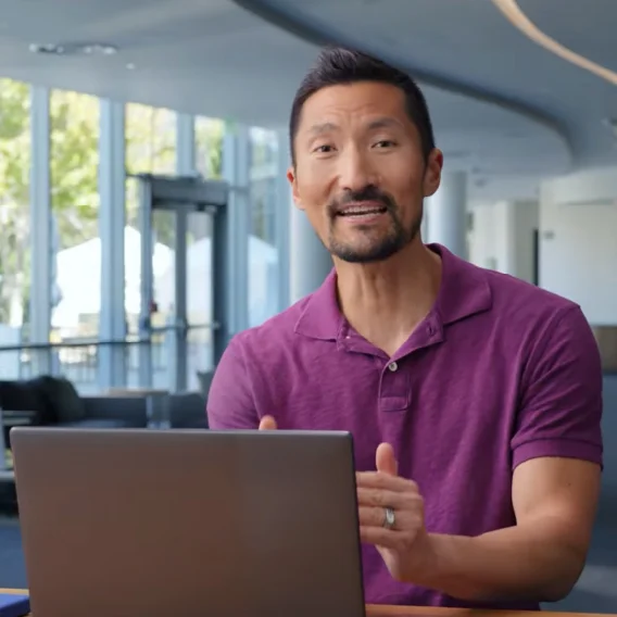 Smiling, bearded man in purple polo gesturing in front of a laptop.